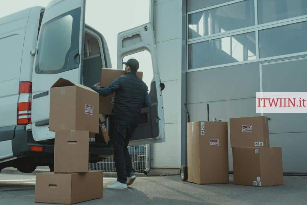A delivery man unloading cardboard boxes from a van at a warehouse during the day.